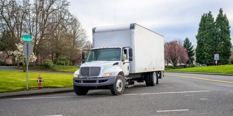 White box truck driving on a street with trees, grass, and a speed limit sign in the background.