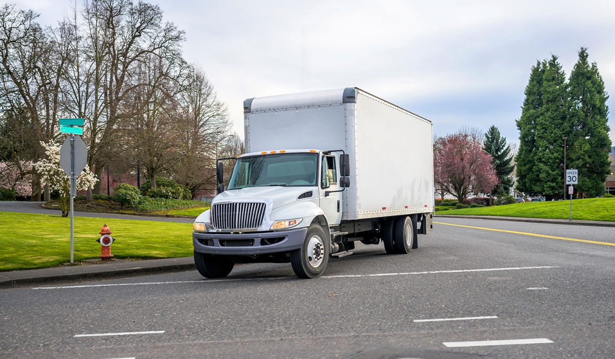 White box truck driving on a street with trees, grass, and a speed limit sign in the background.